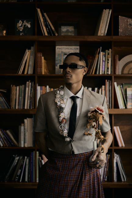 Stylish man in unique attire poses in a modern library with bookshelves.