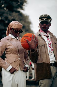 Two adults in vintage attire pose with a basketball outdoors, showcasing retro fashion.