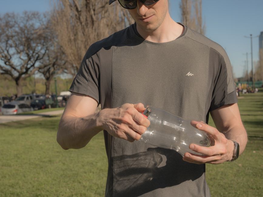 A man in a grey shirt works with a plastic bottle in a sunny park.