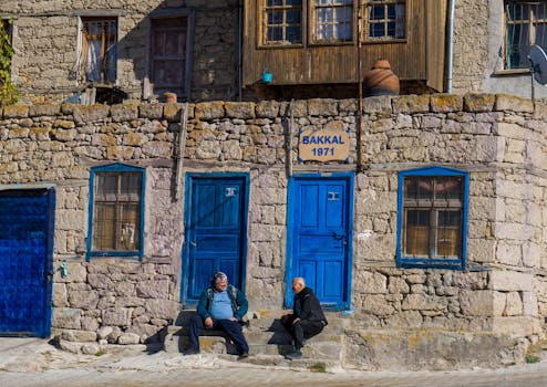 Two men sit outside a rustic stone building with vibrant blue doors in a quaint setting.
