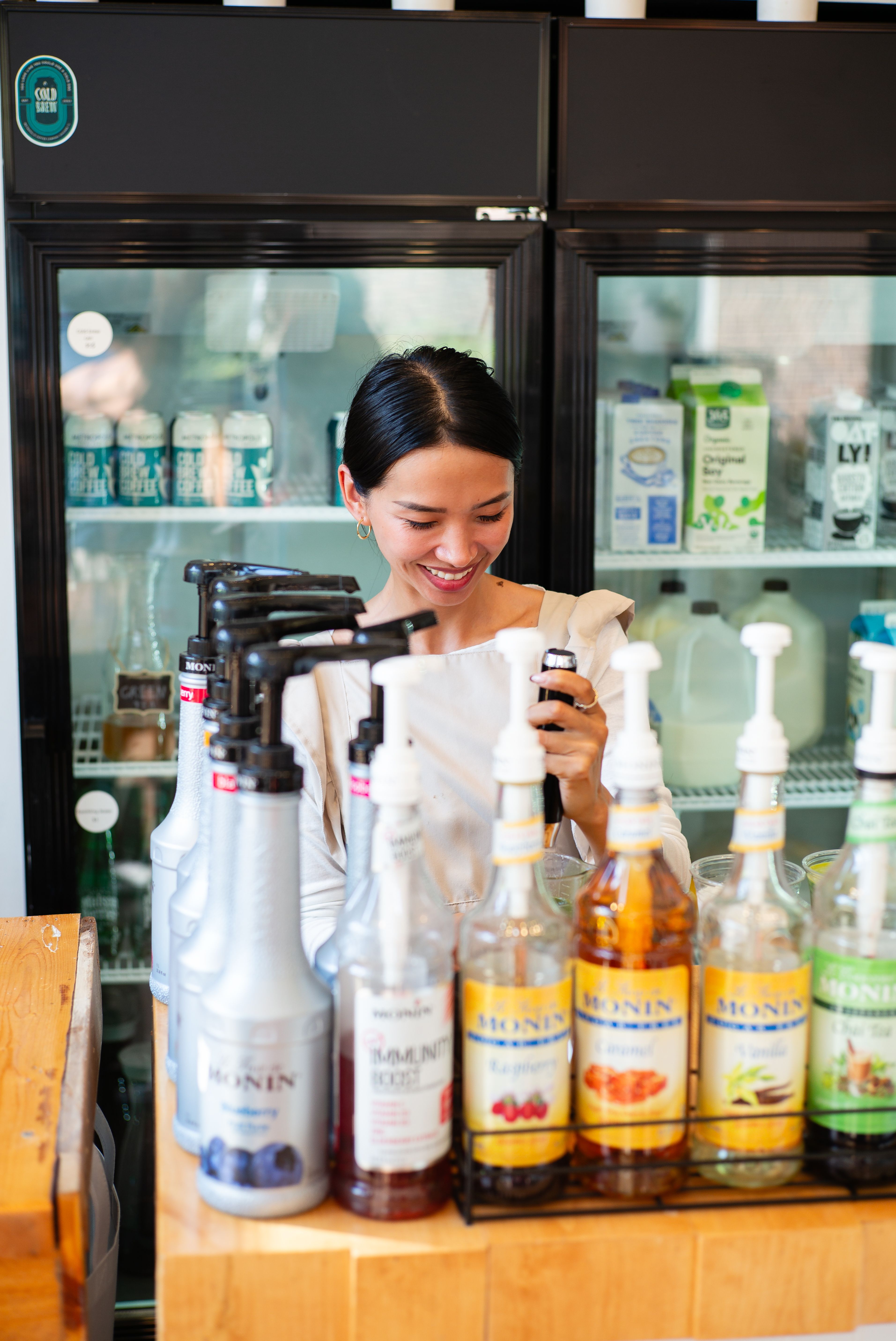 Barista mixes flavors for coffee at a cafe, surrounded by syrup bottles and a refrigerator.