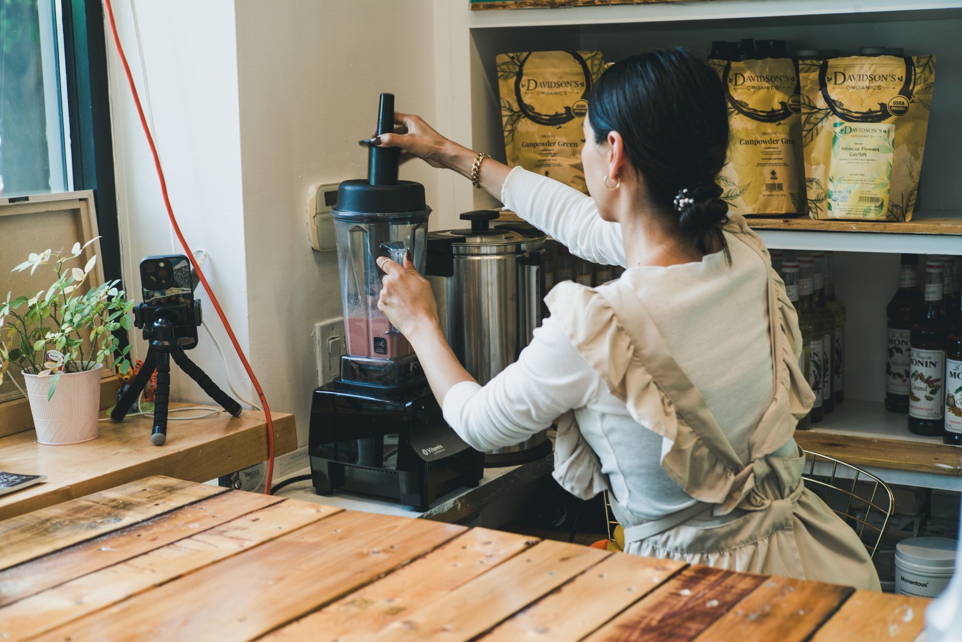 Adult woman making a smoothie in a coffee shop with a blender and camera setup nearby.