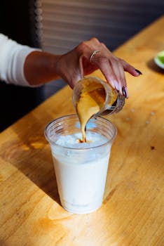 Close-up of a hand pouring espresso into an iced milk drink indoors.