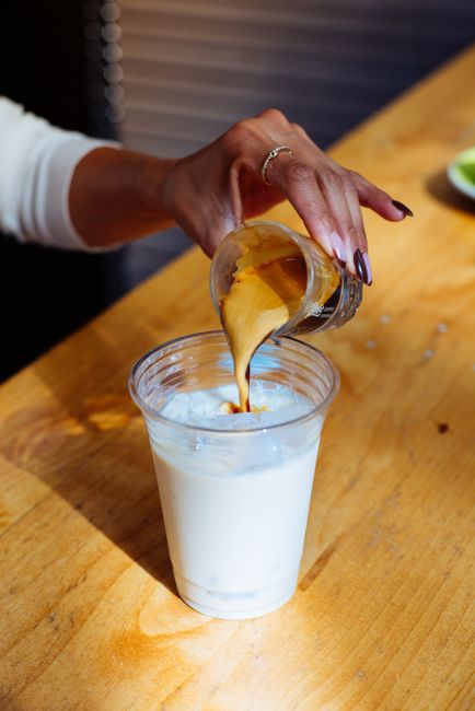 Close-up of a hand pouring espresso into an iced milk drink indoors.