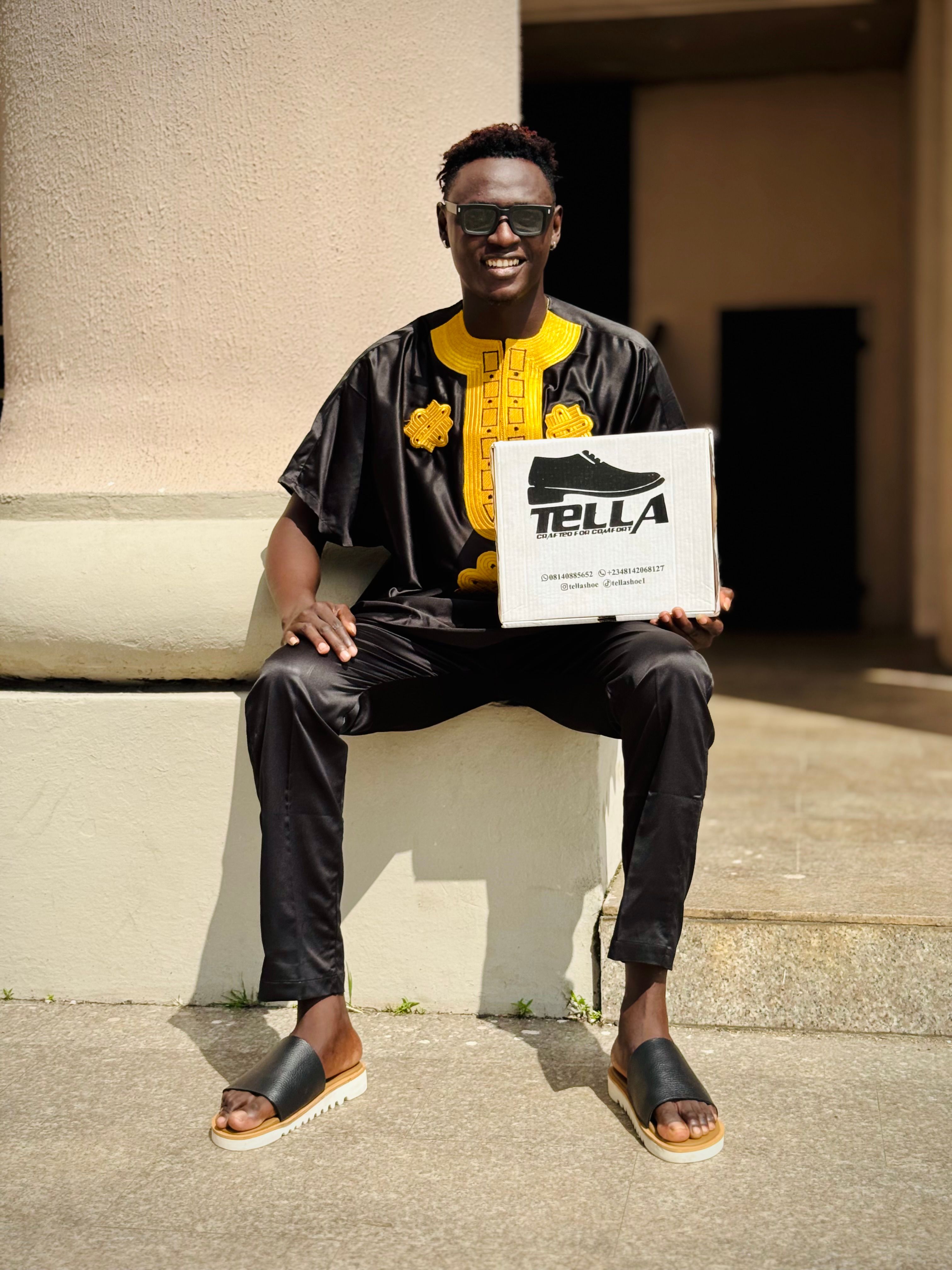 Cheerful man in African attire holding Tella shoebox, sitting outside.
