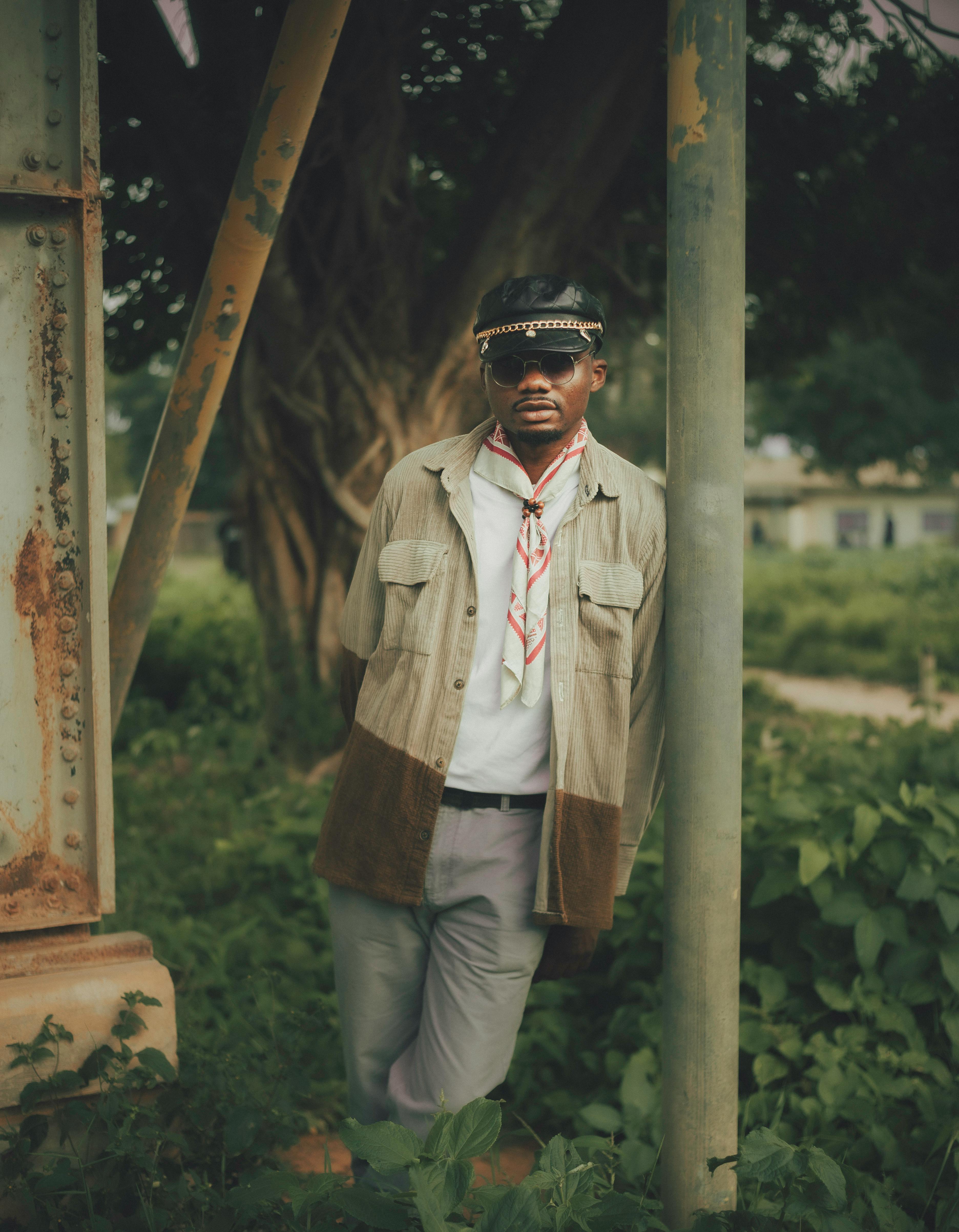 Man in casual vintage clothing outdoors, leaning on rustic metal beam surrounded by greenery.