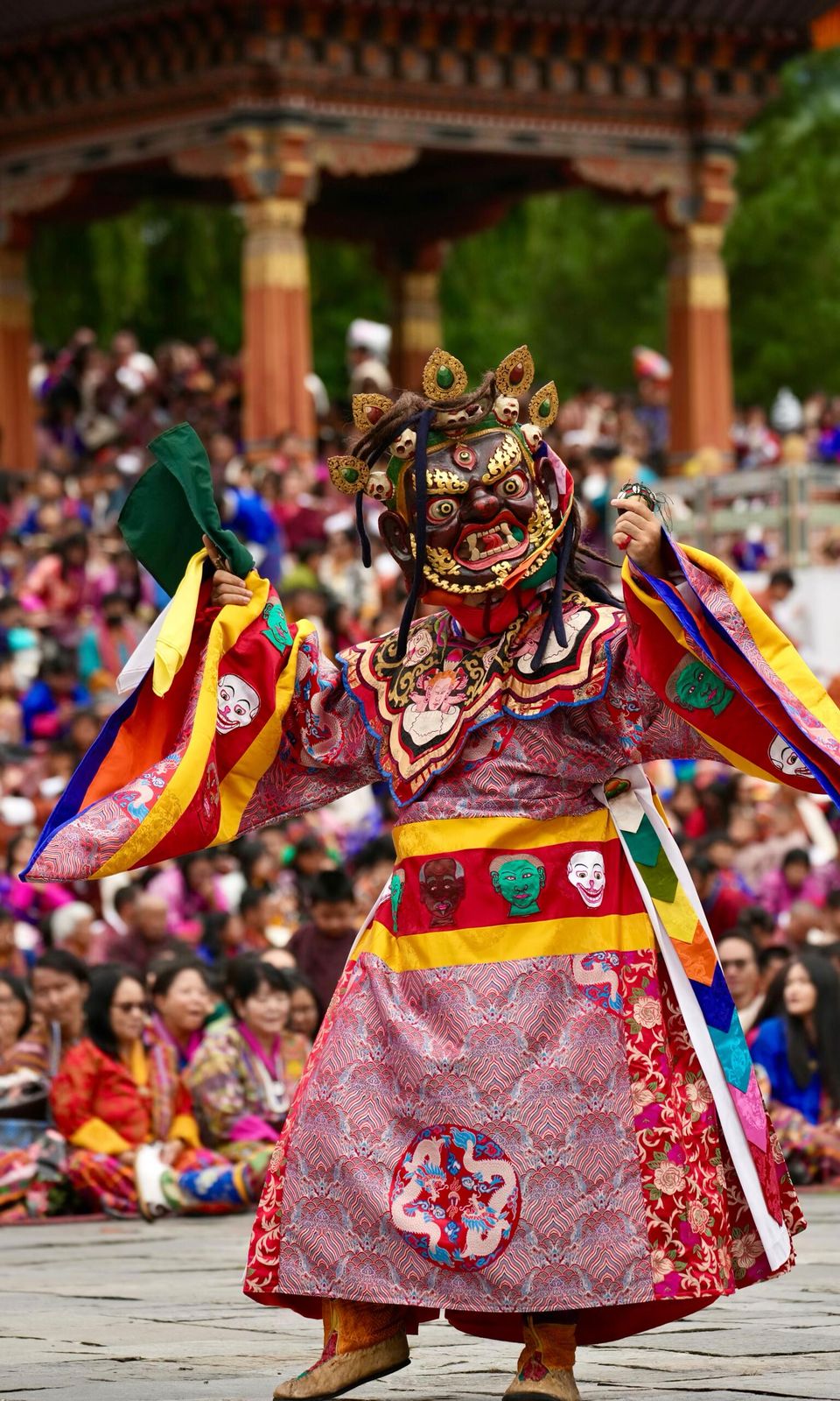 A masked cham dancer in brocade and silk at a tshechu in Thimphu, Bhutan, the seated crowd behind