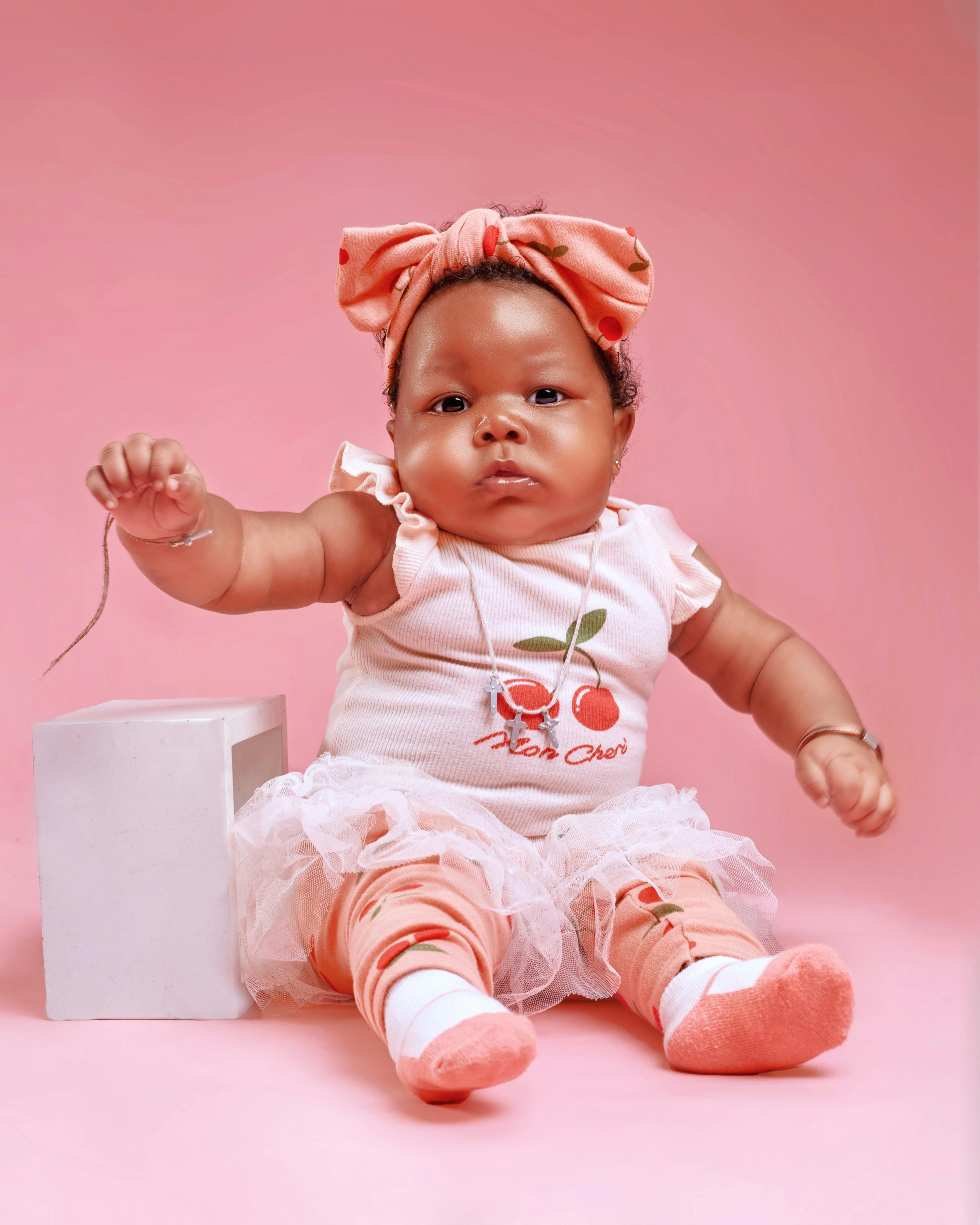 Charming baby in pink attire with cherry motif, sitting against a soft pink backdrop.