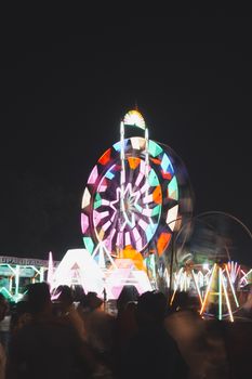 Colorful Ferris wheel lights up night sky at a lively carnival, surrounded by blurred crowds.