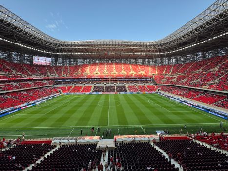 A vibrant football stadium with a broad green field and red seats filled with sports fans.