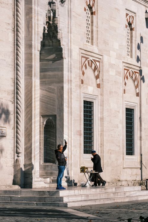 Free A street musician plays an instrument outside a historic building while a person takes a photo. Stock Photo