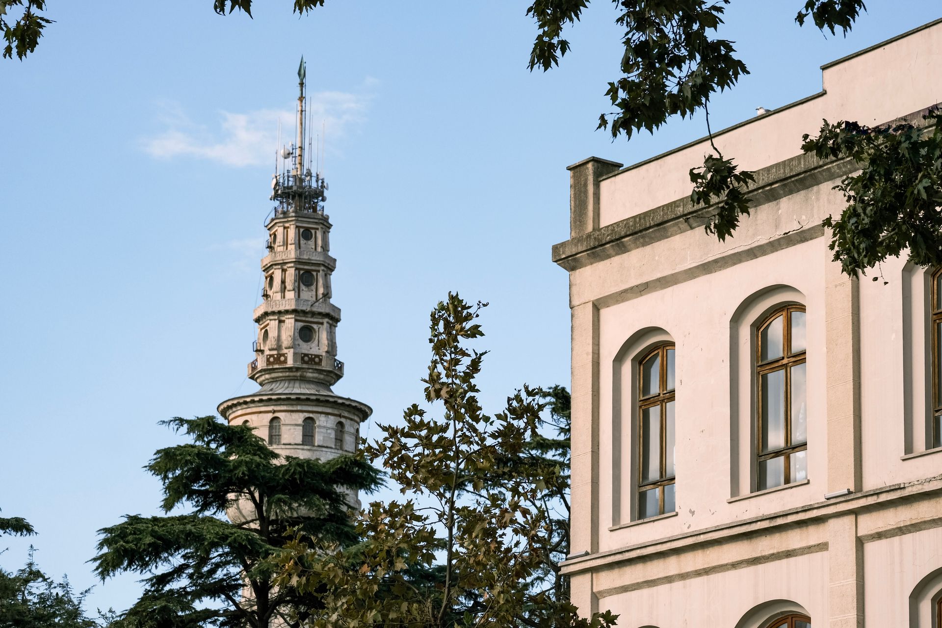 A historic tower and adjacent building surrounded by lush trees under a clear blue sky.