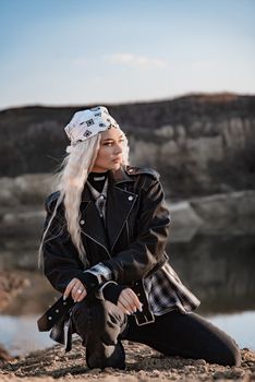 Young woman wearing a leather jacket and bandana, kneeling outdoors by water.