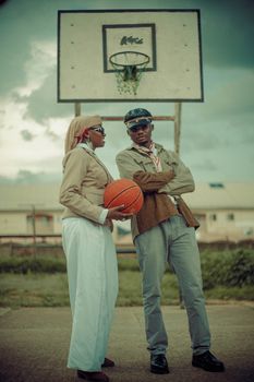 Two fashionable adults pose with basketball on outdoor court.