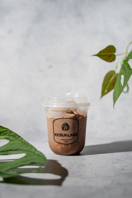 Free Refreshing iced chocolate drink in a branded plastic cup surrounded by lush green leaves on a concrete surface. Stock Photo