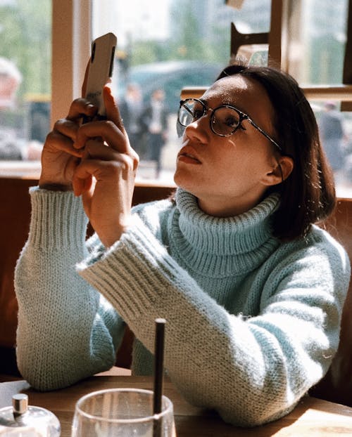 Woman Wearing Light-green Turtleneck Sweater and Eyeglasses Using Phone While Sitting Near Brown Wooden Table Beside Glass Wall