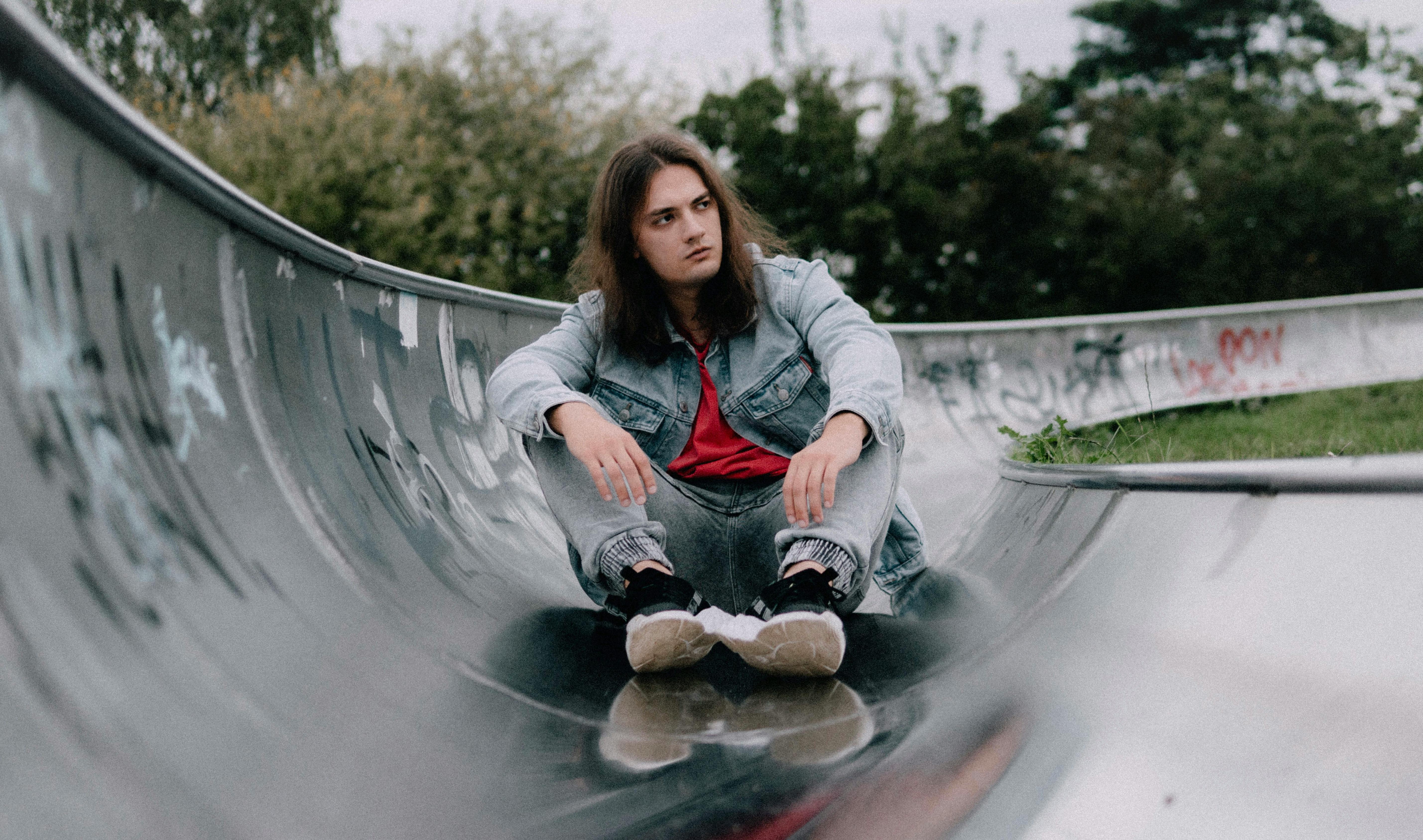 Gratis Un giovane con una giacca di jeans è seduto in uno skatepark, in atteggiamento riflessivo. Foto a disposizione