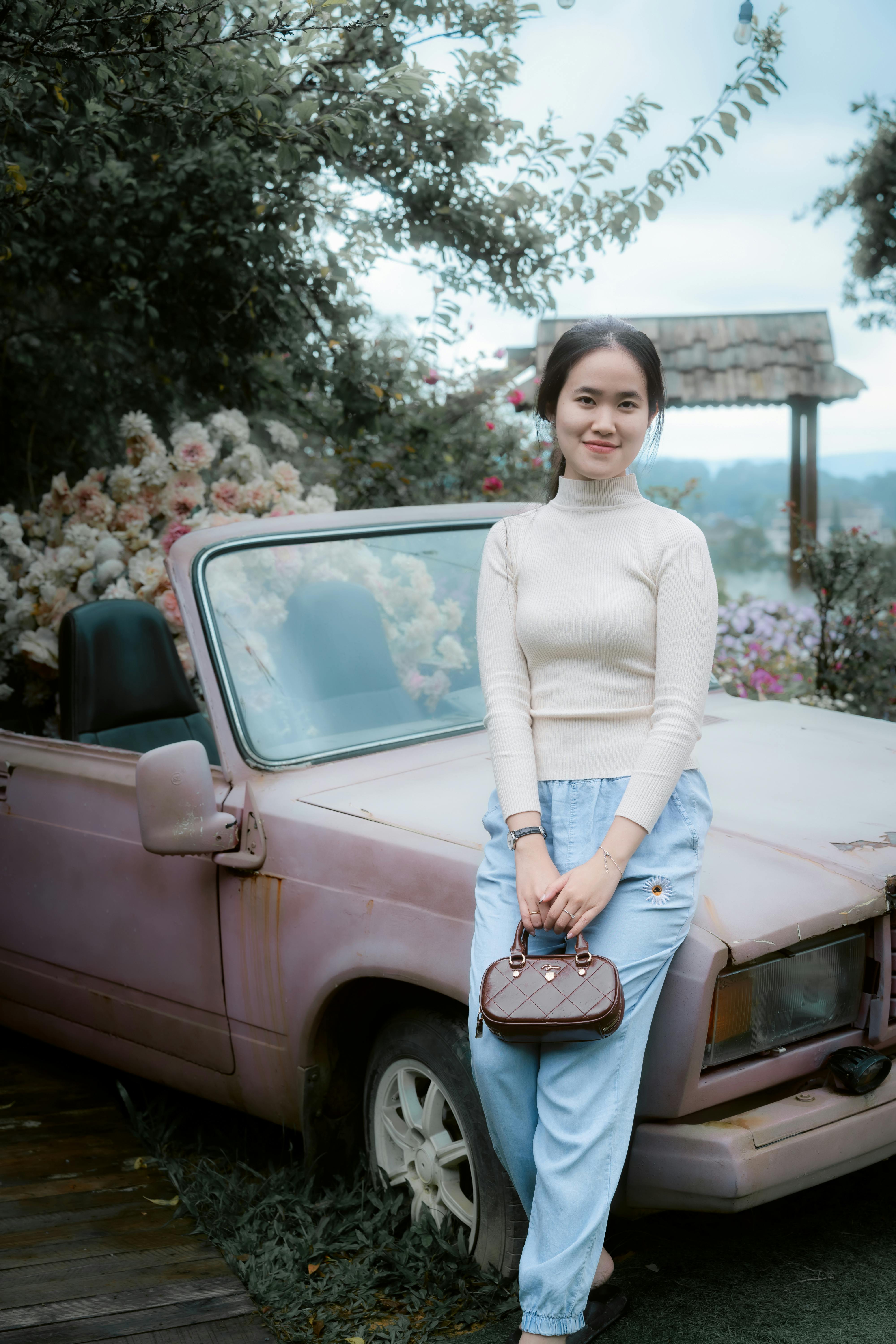A young woman stands by an old vintage car surrounded by flowers in a serene outdoor garden setting.