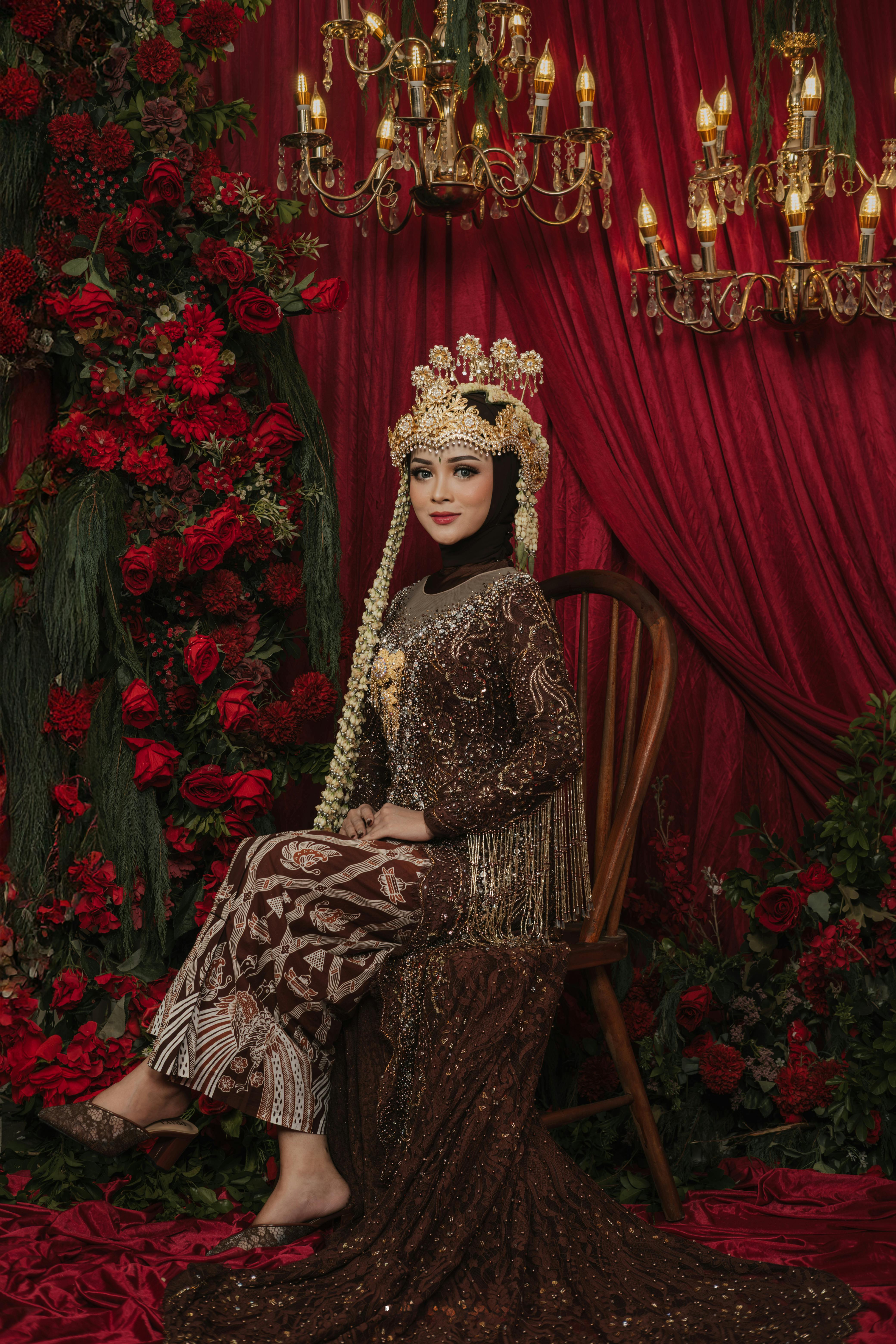 Indonesian bride in ornate traditional attire with a backdrop of chandeliers and red flowers.