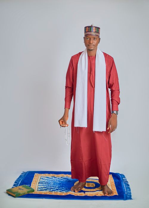 Free Man wearing traditional red outfit and cap standing on a blue prayer mat indoors. Stock Photo