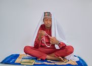 A young man in traditional attire sits cross-legged with prayer beads and a Quran, showing a moment of spiritual reflection.