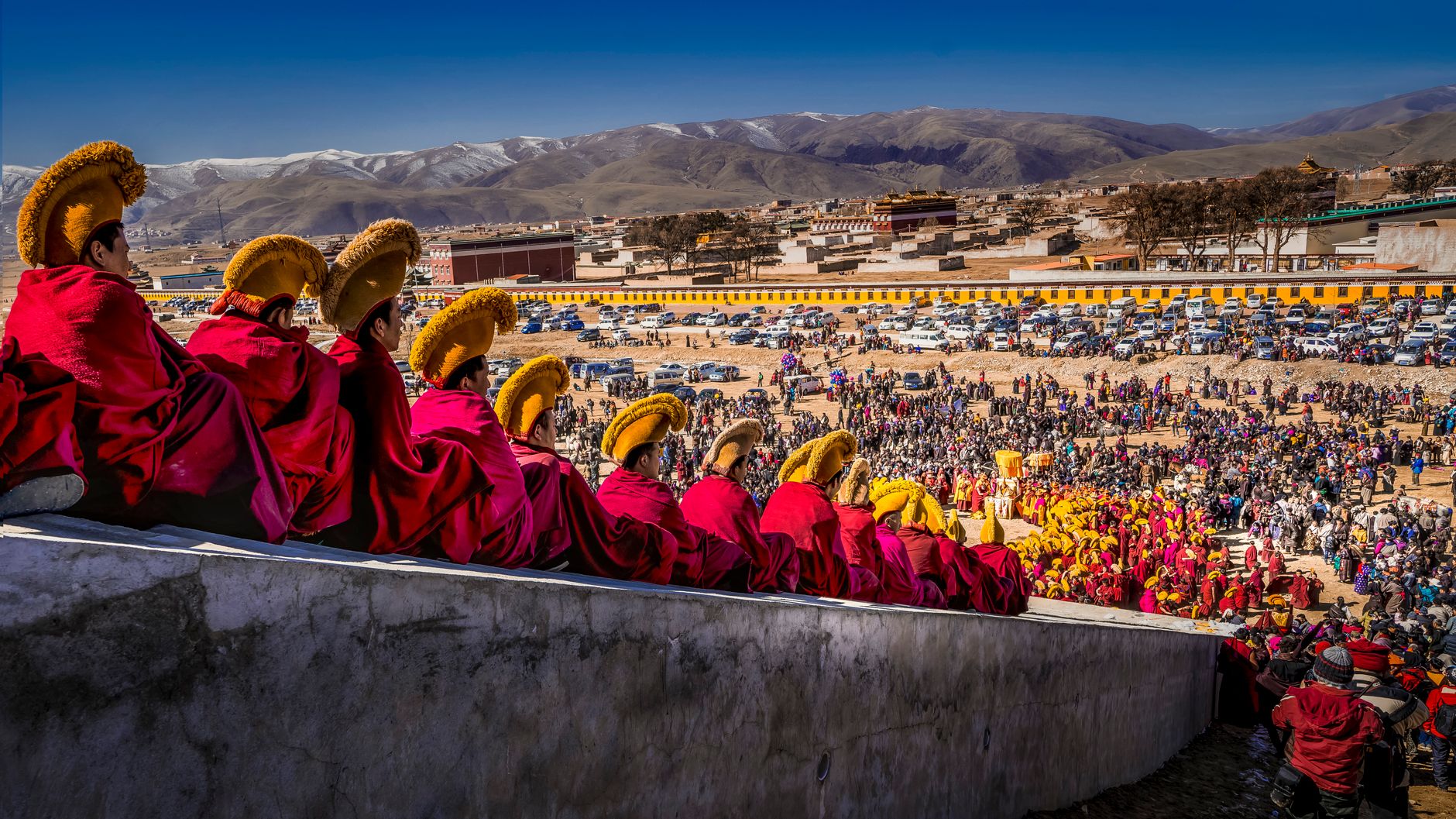 Tibetan monks in traditional attire observe a vibrant festival in a mountainous setting.