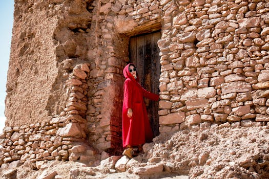 Woman in a red coat standing by a rustic stone wall under bright sunlight.