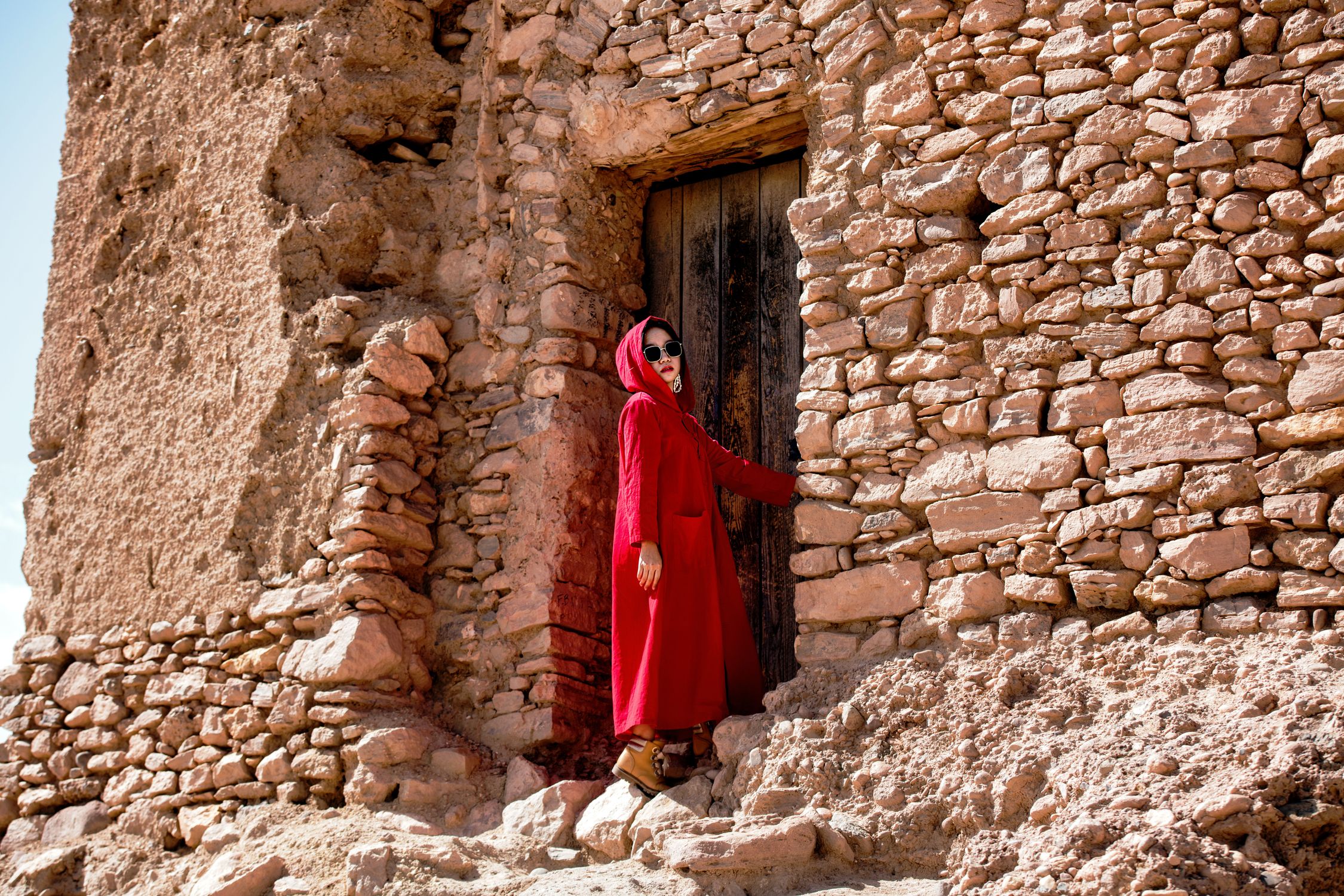 Woman in a red coat standing by a rustic stone wall under bright sunlight.