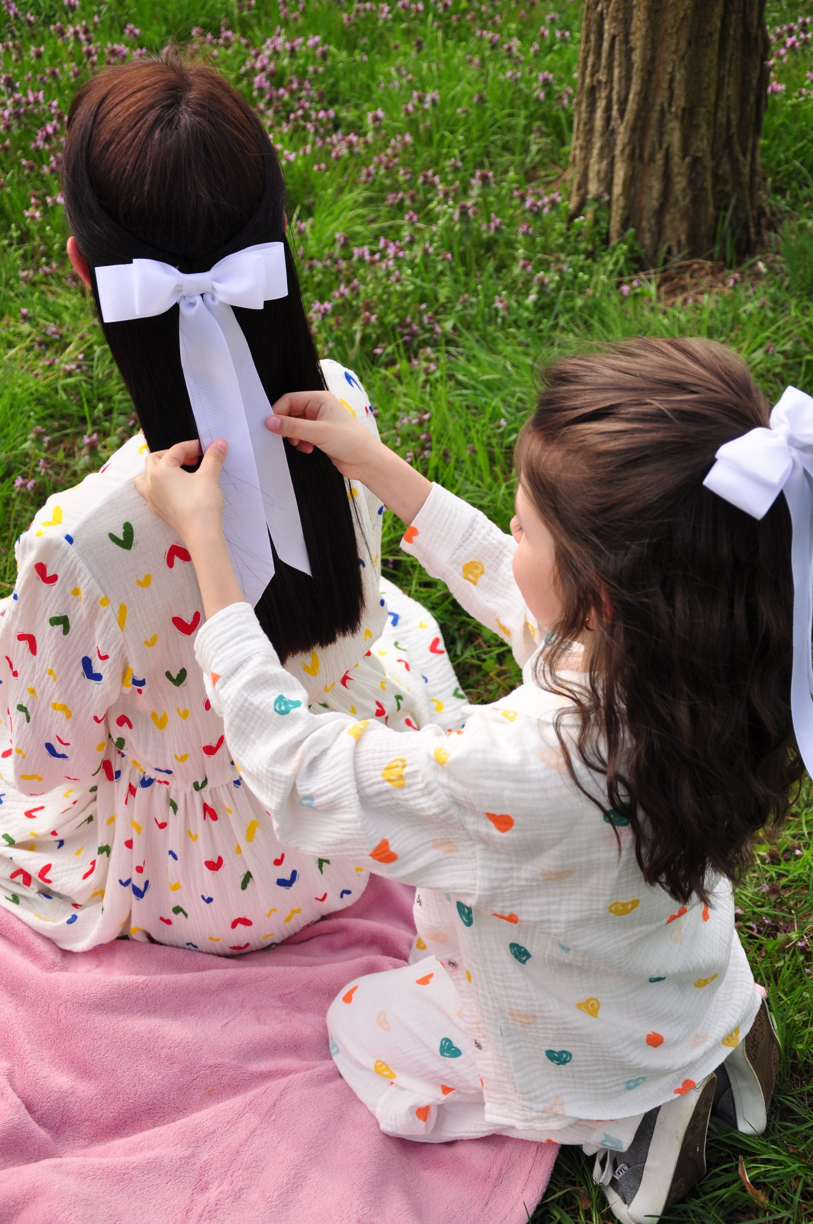 Two sisters wearing colorful outfits, sitting on grass, tying hair bows.