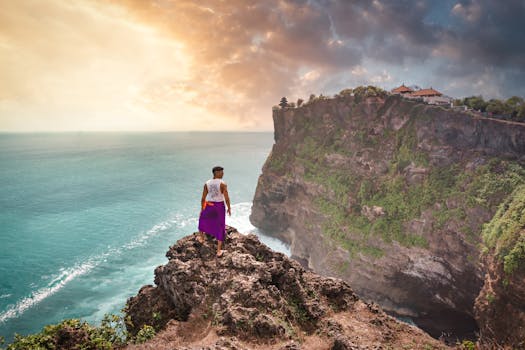 Man standing on cliff with scenic view of Uluwatu Temple, Bali at sunset.