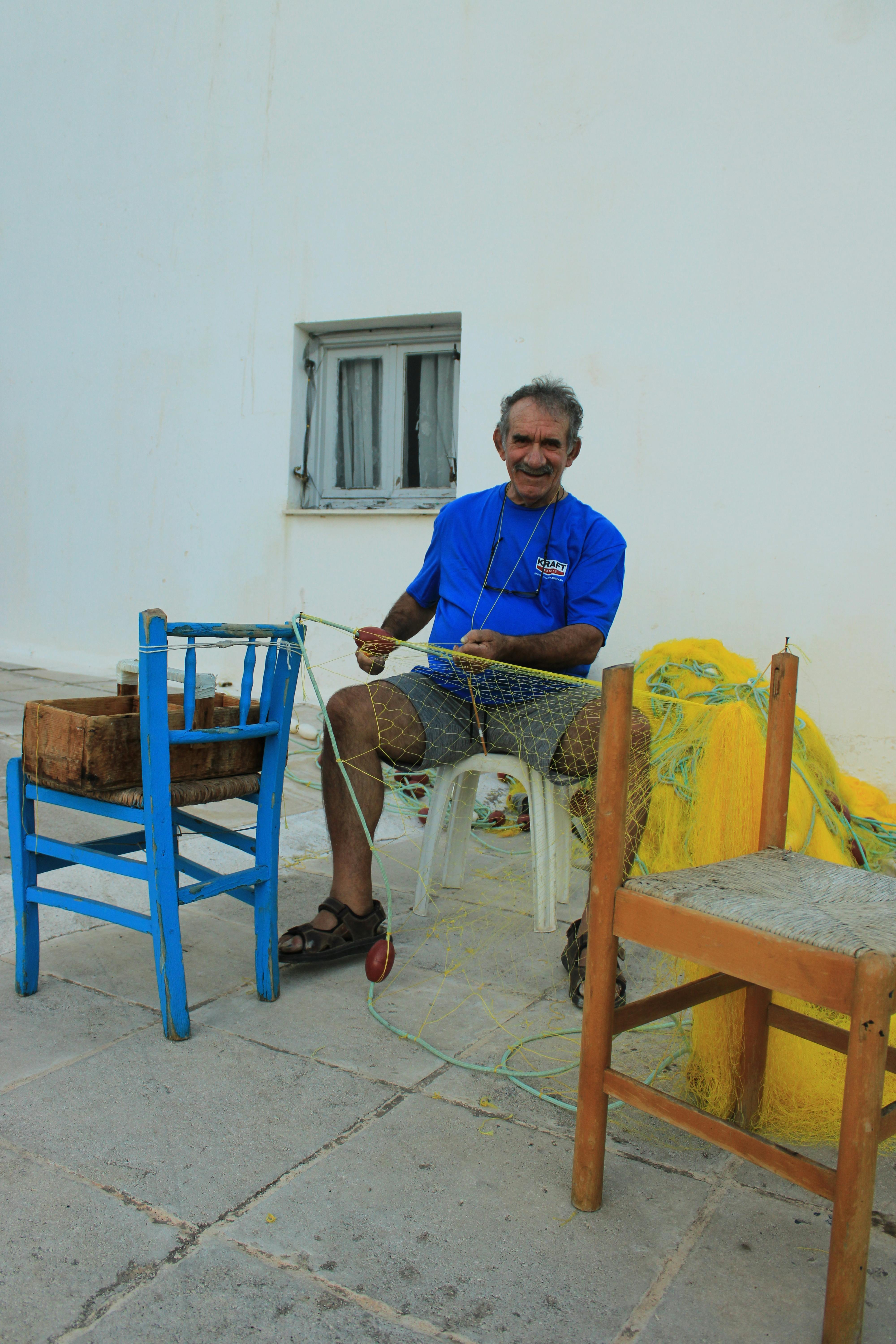 An elderly fisherman mending a fishing net outside against a white wall.