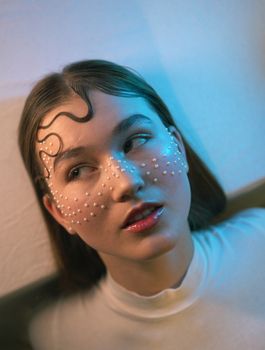 A woman with artistic pearl makeup under soft, colorful lighting.