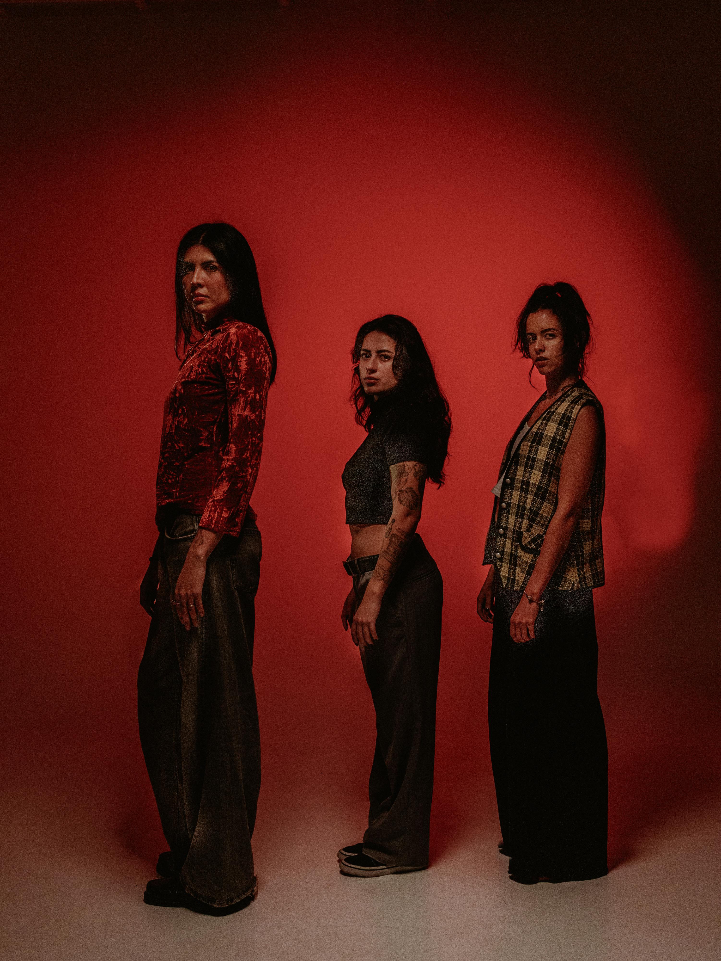 Three diverse women pose in a moody, red-lit studio, exuding confidence and style.