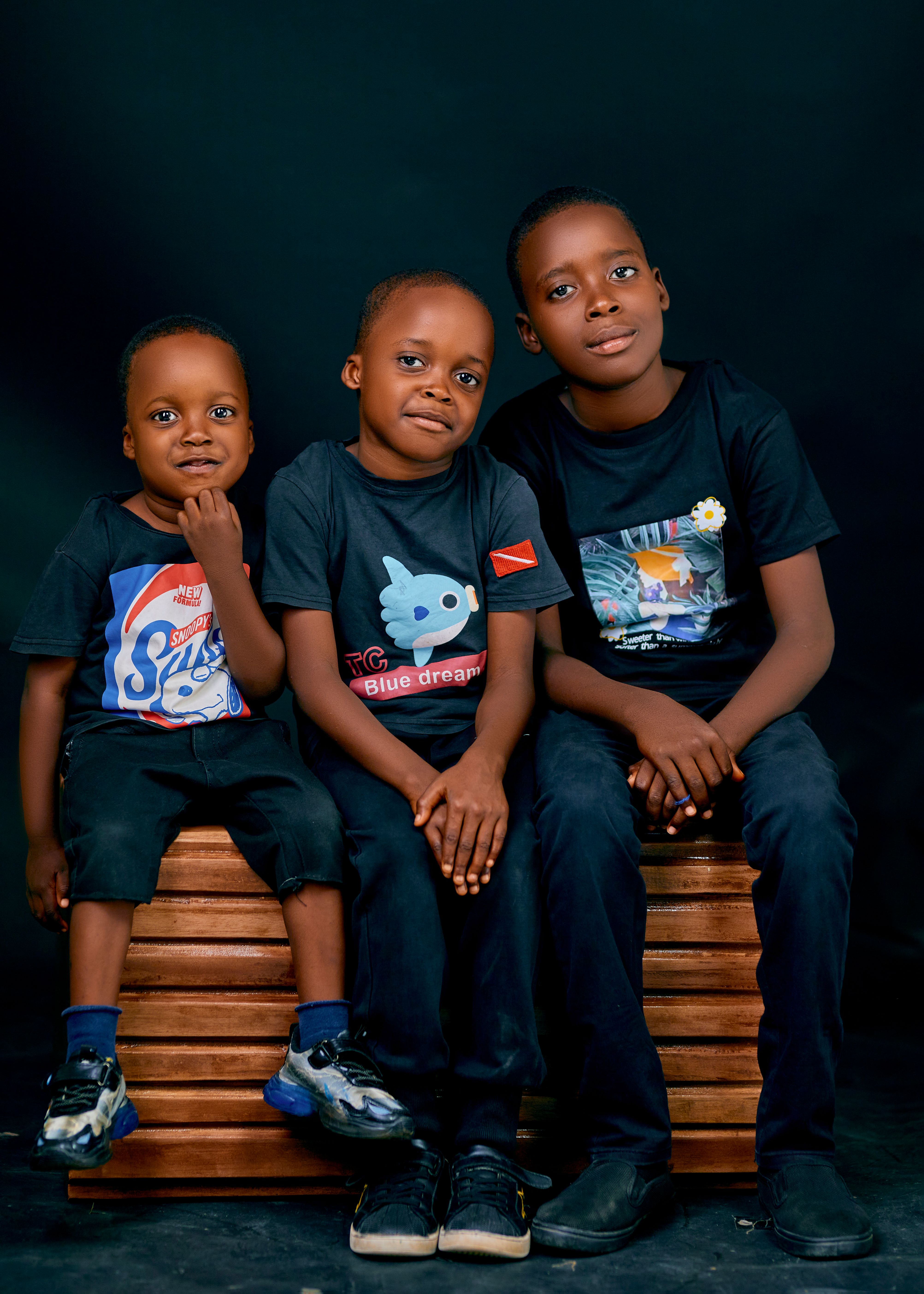 Three smiling children sitting on a bench against dark background, wearing casual outfits.