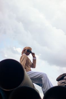 Fashionable person in sunglasses and headscarf posing on industrial pipes with cloudy sky.