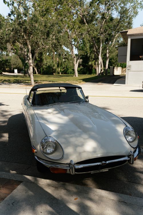 Free A vintage white convertible parked on a sunny street beneath leafy trees. Stock Photo