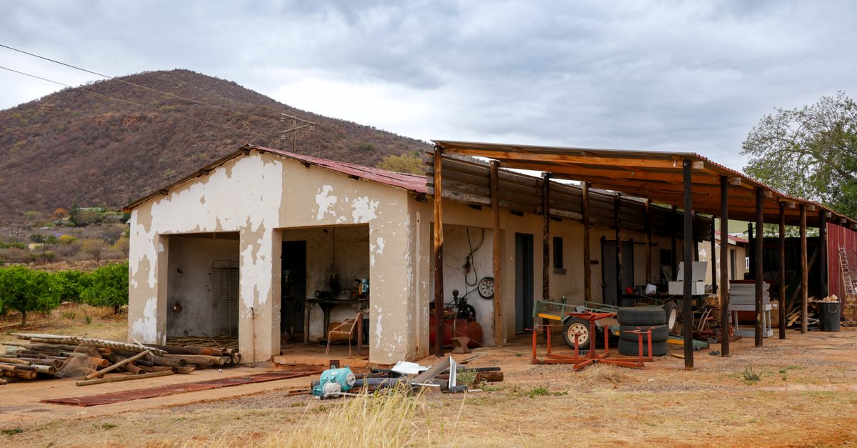 Rustic farmyard with a workshop and mountains in the background under cloudy skies.