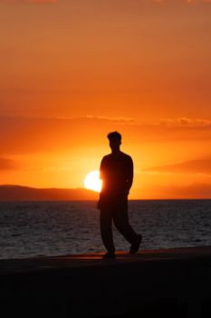 Silhouette of a person walking along the shore during a vibrant sunset over the ocean.
