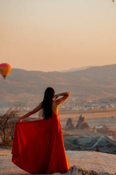 A woman in a flowing red dress enjoys the stunning sunrise view of Cappadocia with hot air balloons in the background.
