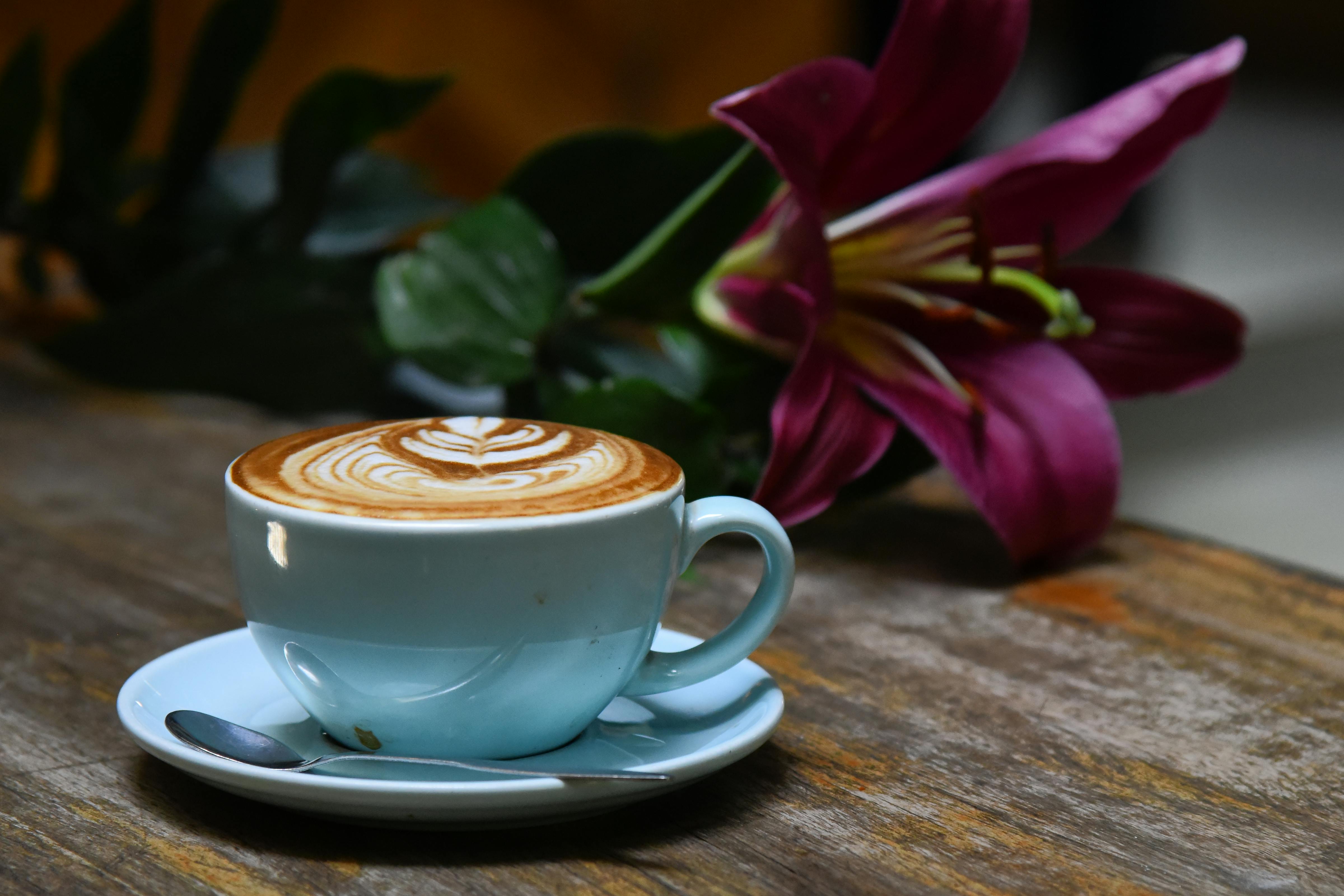 A beautifully crafted latte with intricate art beside a vibrant purple lily on a rustic wooden table.