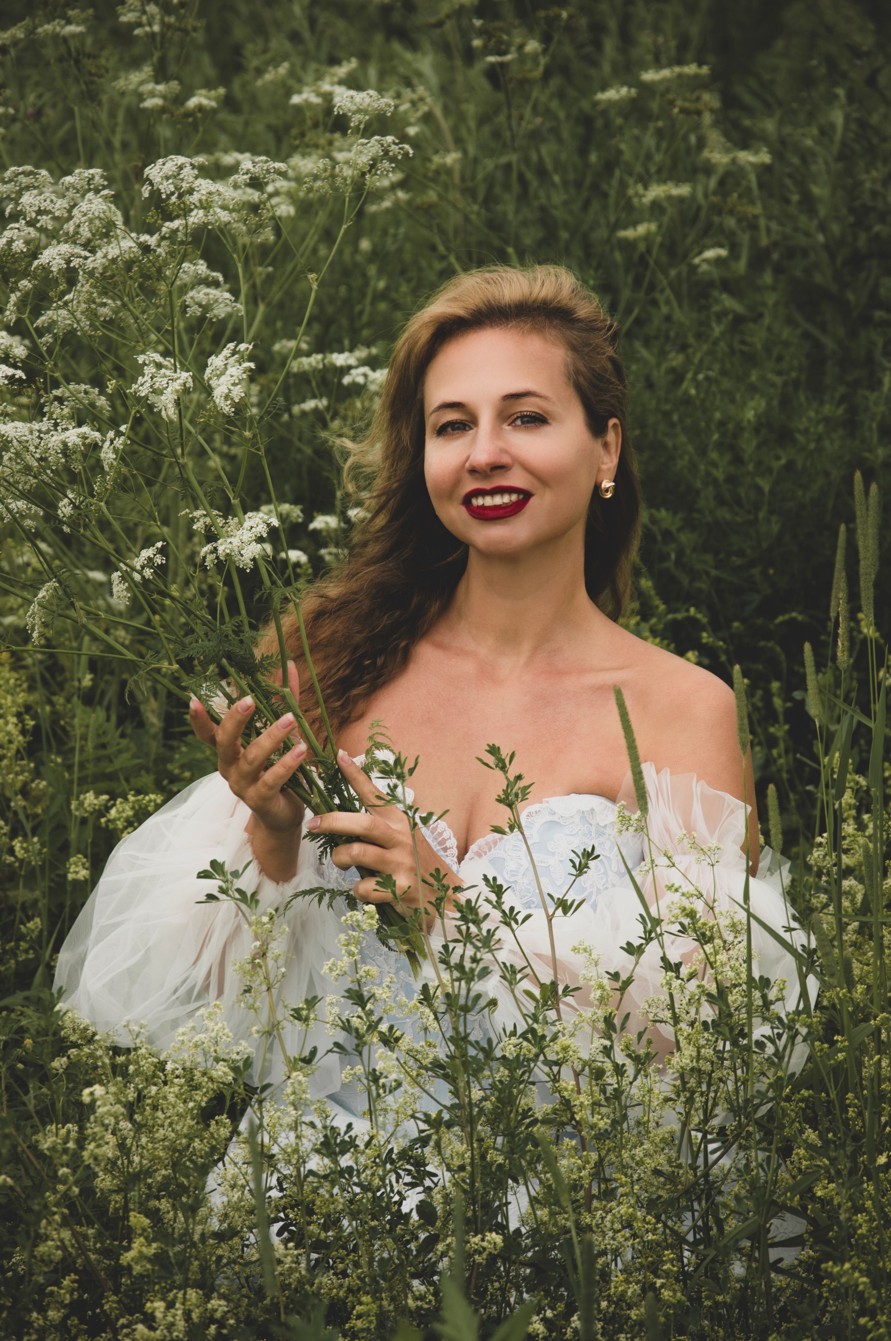 A woman in a white dress smiles amid tall wildflowers in a lush green field.