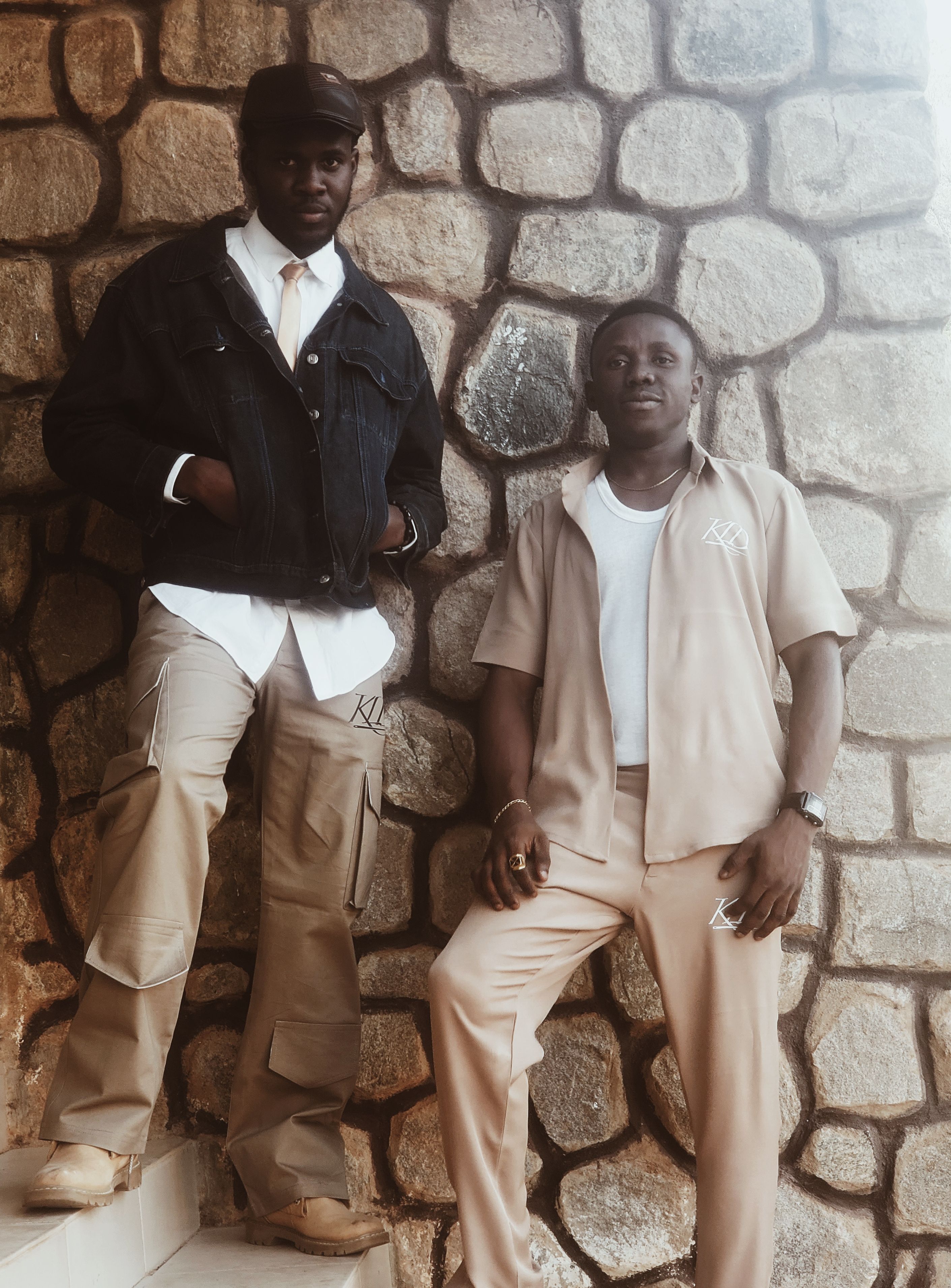 Two stylish men stand confidently in front of a stone wall, showcasing contemporary fashion.
