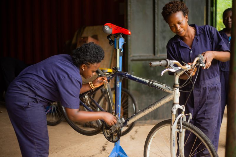 Two female mechanics in blue overalls working on a bicycle inside a workshop.