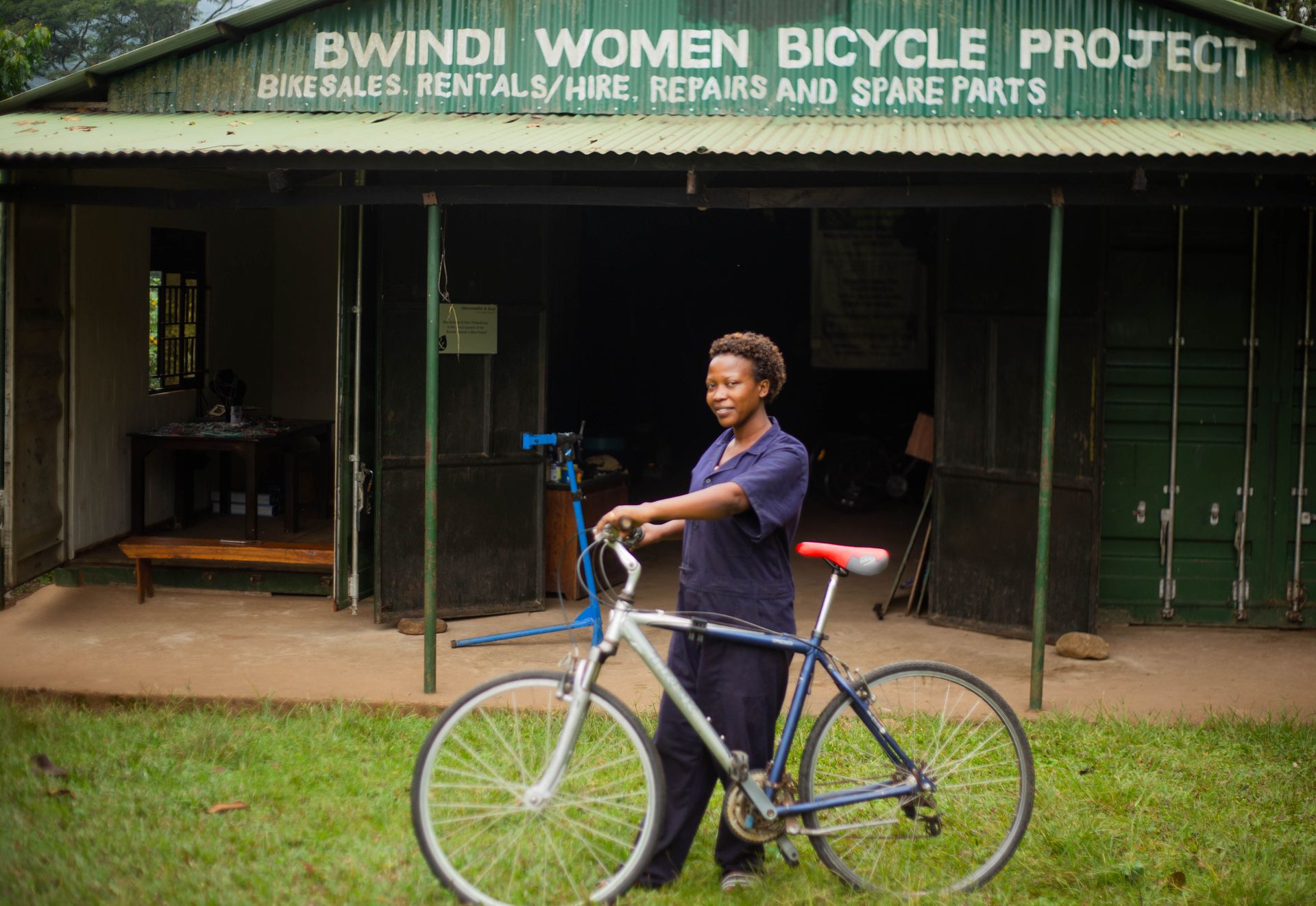 Smiling woman holding a bicycle outside Bwindi Women Bicycle Project shop. Empowerment and community support theme.