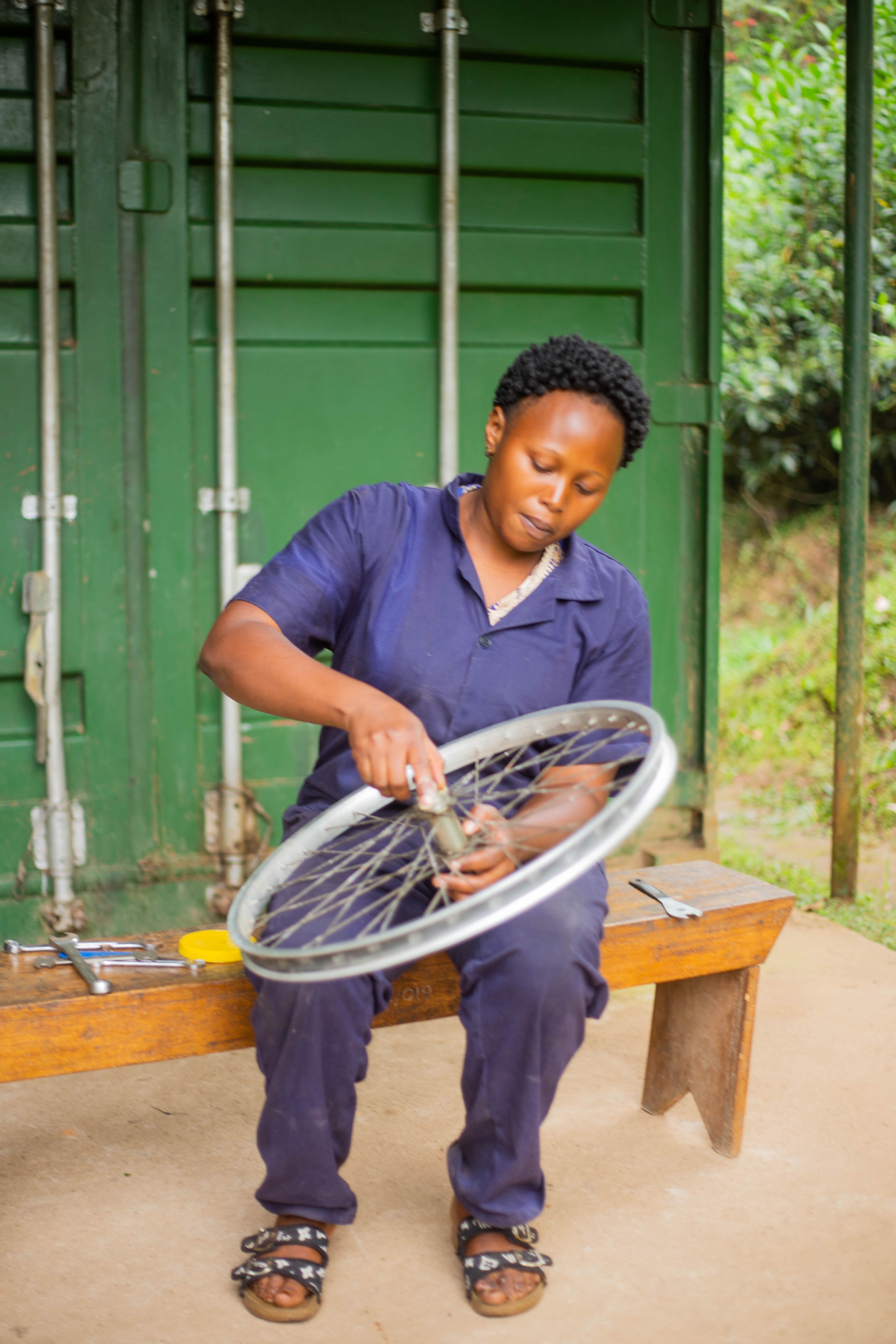 A woman in overalls working on a bicycle wheel outside with tools on a bench.
