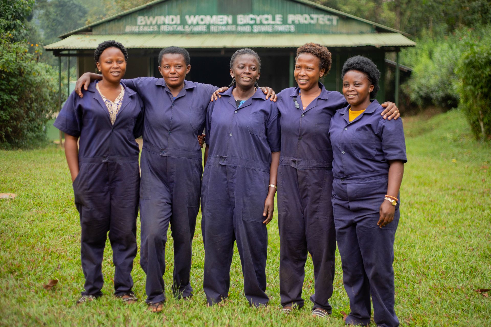 Five women in overalls at Bwindi Women Bicycle Project show empowerment and teamwork.
