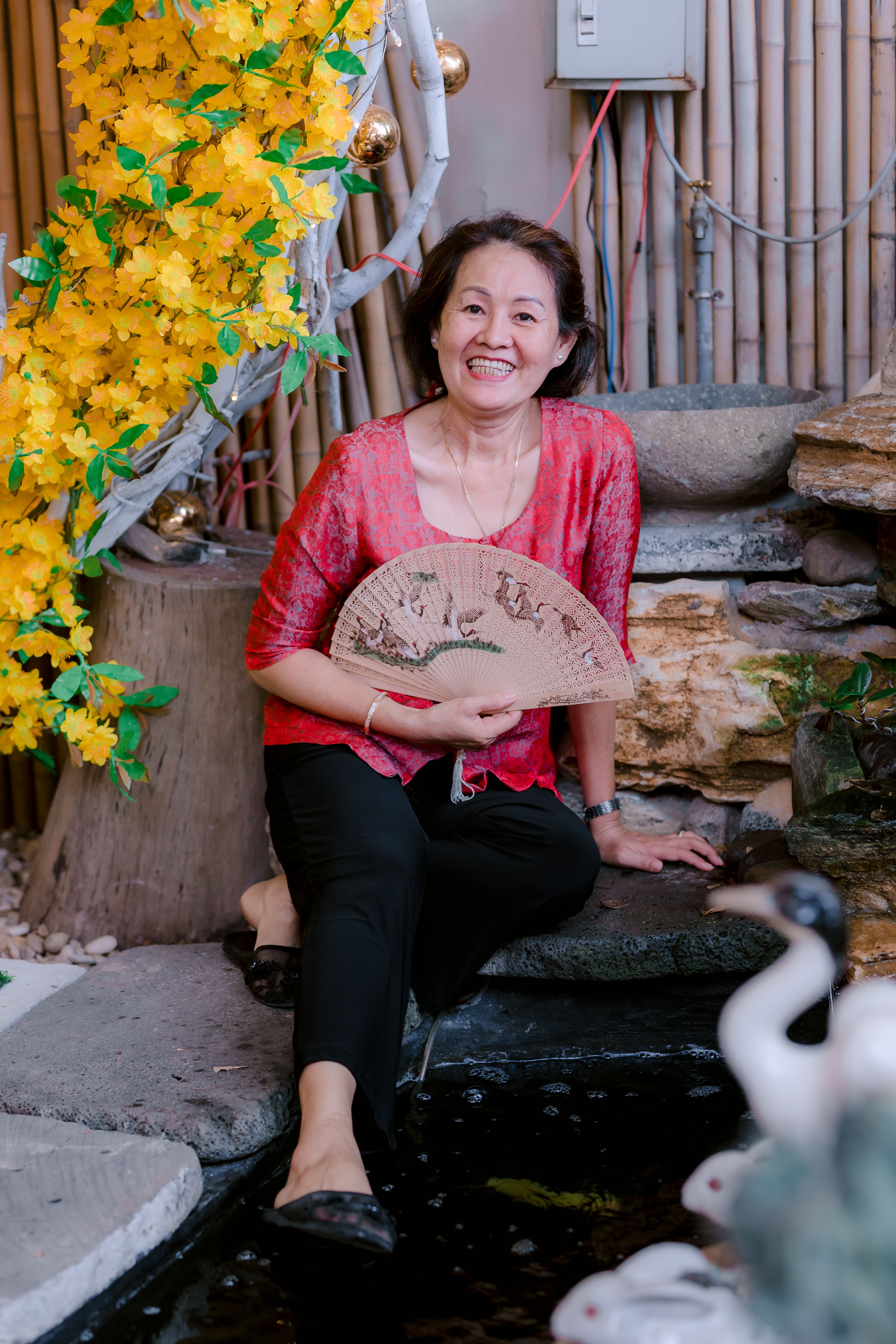 A cheerful woman holding a fan, sitting in a serene garden with vibrant flowers.