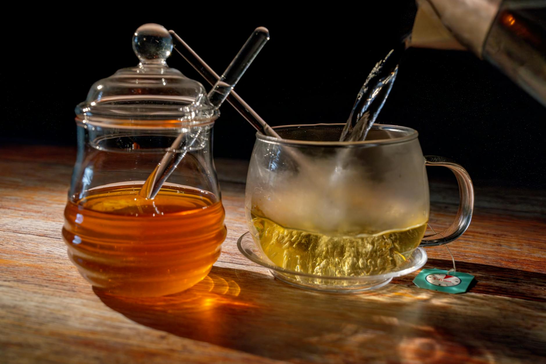 Ahşap bir zemin üzerinde buharı tüten bir fincan ıhlamur çayı, yanında bal kavanozu ve kurutulmuş ıhlamur çiçekleri.