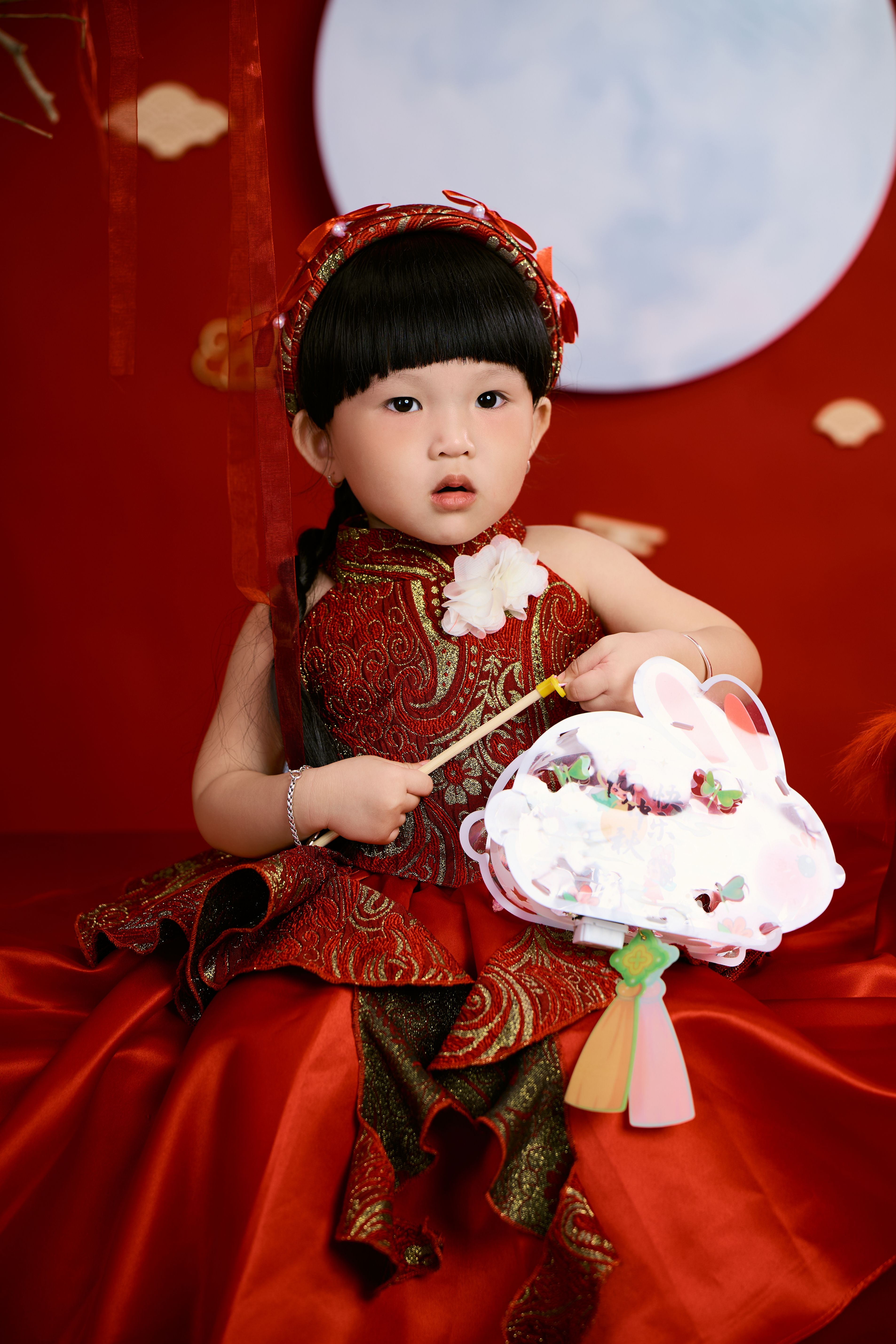 Cute child wearing traditional red dress holding decorative fan against red backdrop.