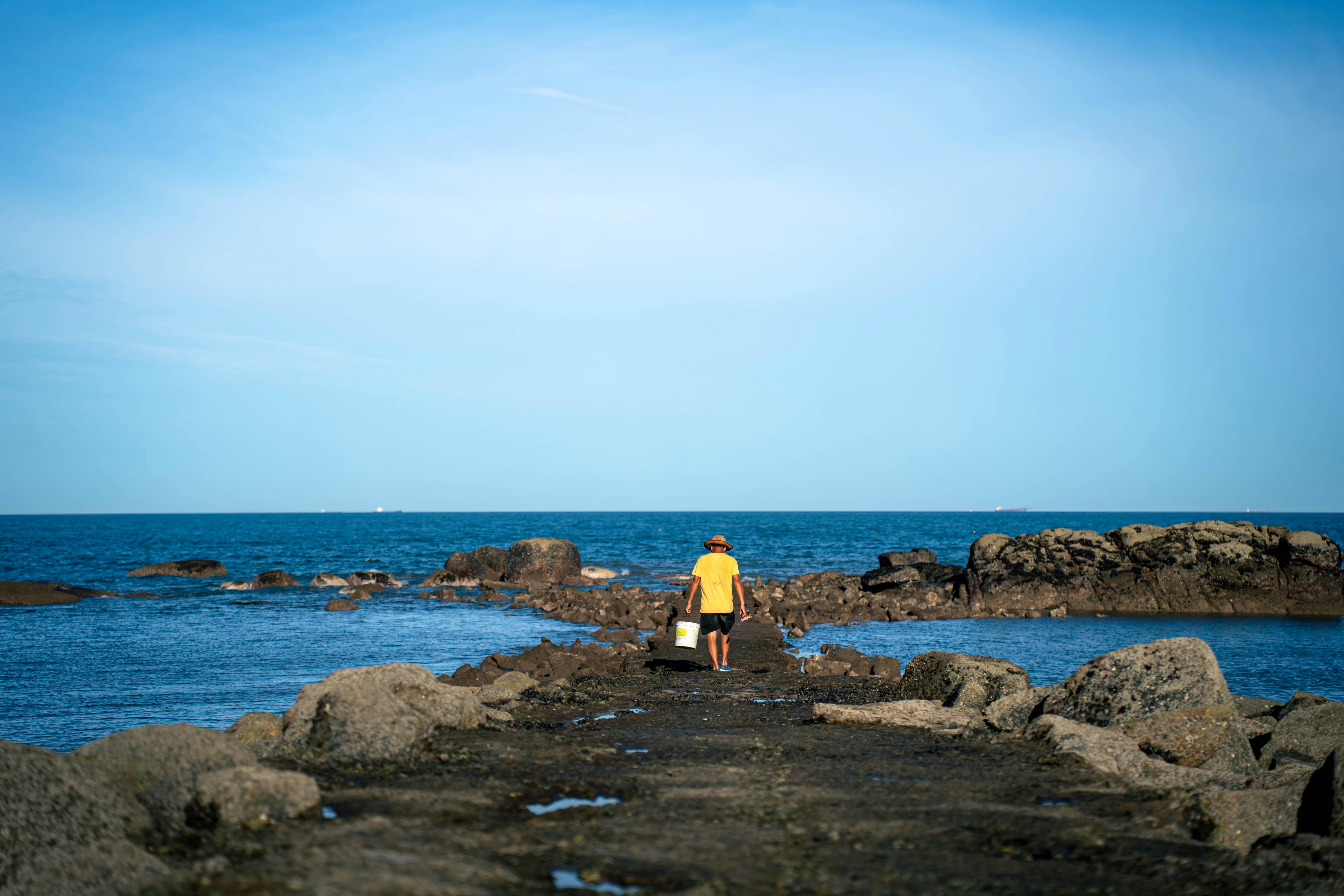 在晴朗的蓝天下，一名渔民沿着岩石海滩走向大海